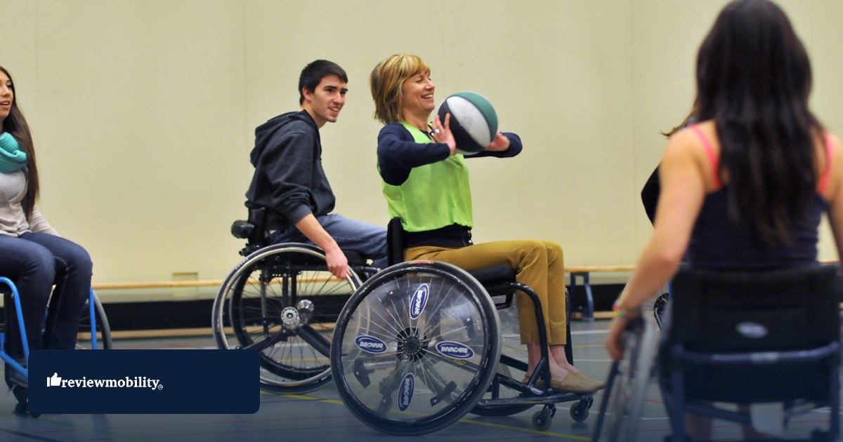 Wheelchair basketball players during a match Group of people in wheelchairs playing basketball in a gym; woman in a green vest throws a ball, others watch; reviewmobility logo visible on lower left.