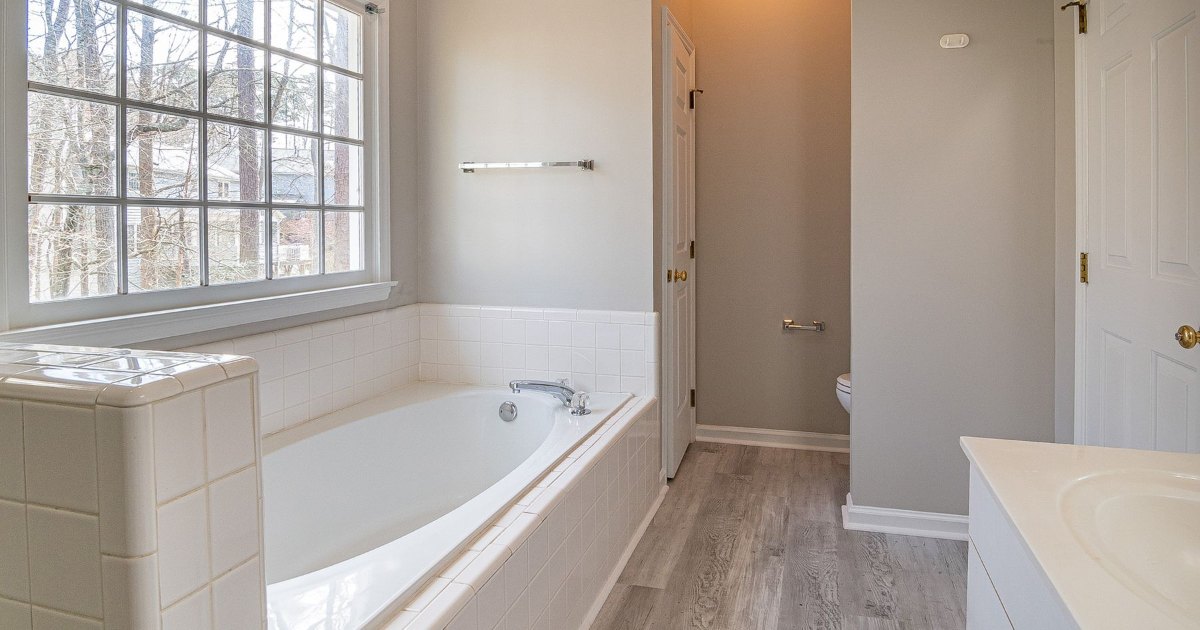 A bath set beneath a window in a bathroom with grey wood effect flooring