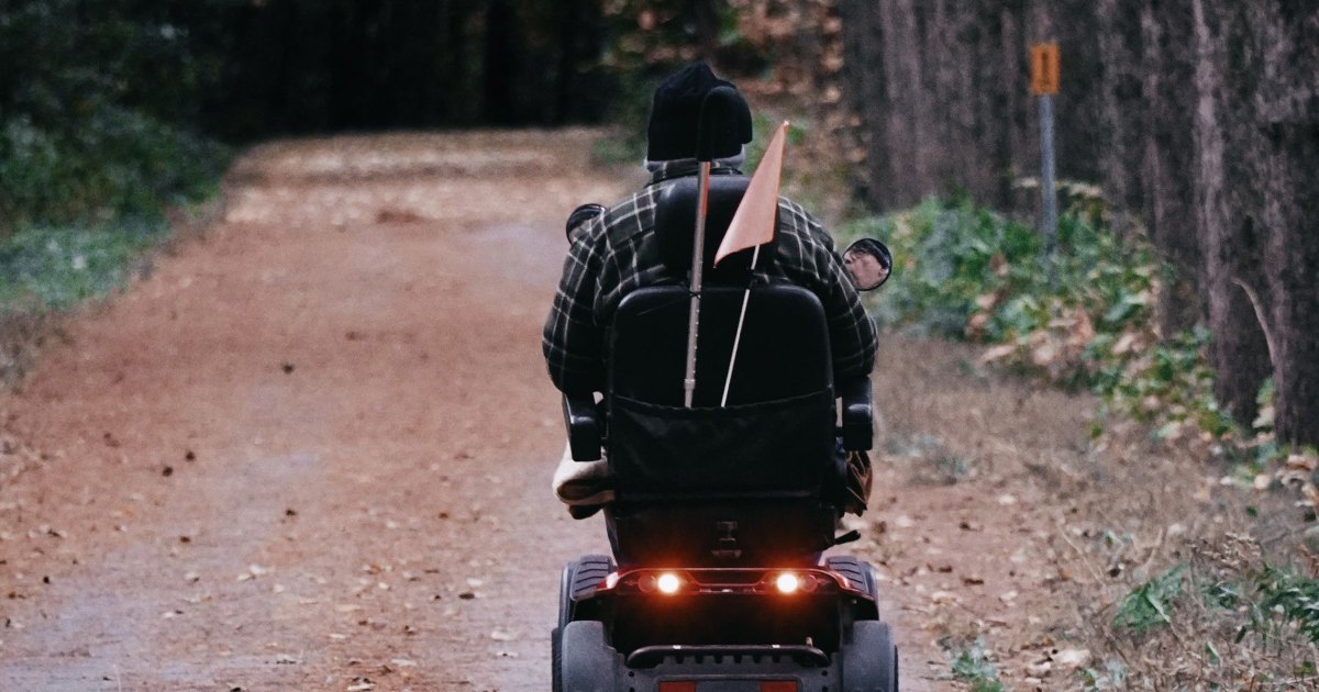 A person riding a mobility scooter along a path through woodland, seen from behind