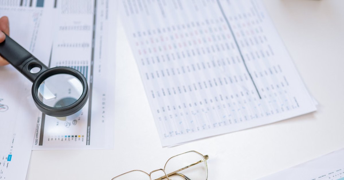 Desk covered with printed documents and charts, a magnifying glass and reading glasses laid out for checking paperwork.